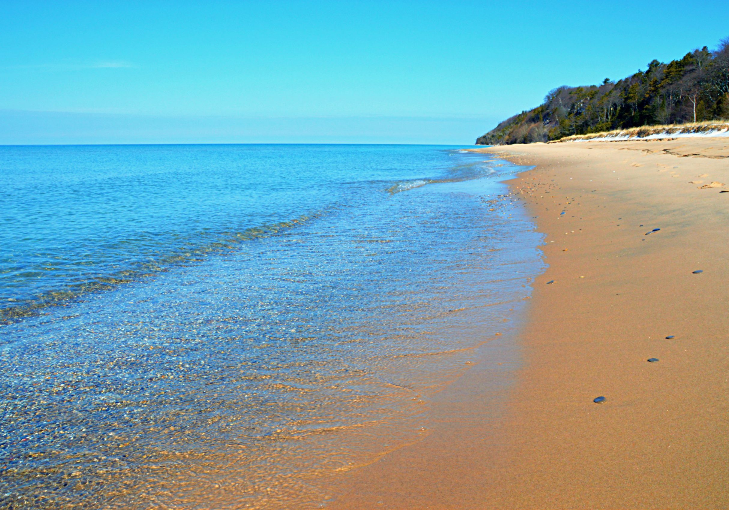 A view of calm water, colorful stones, and sandy beach at North Beach in Leland, Michigan, along the shores of Lake Michigan on a sunny, warmer-than-normal, February afternoon.